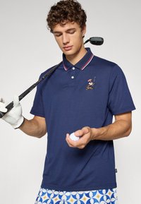 Tamsiai mėlynas polo marškinėlių su raudonai baltais dryžuotais apykakle, su siuvinėtu personažu. Modelis laiko golfą kamuoliuką ir lazdą, vilkėdamas raštuotas šortus.