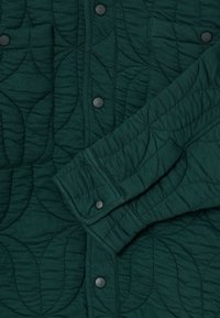 Σκούρο πράσινο καπιτονέ μπουφάν με κυκλικό σχέδιο ραφής, κουμπιά με γατζάκια και μακριά μανίκια που είναι εν μέρει διπλωμένα μπροστά.