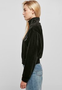 Chaqueta corta de terciopelo negro con cuello elevado, puños acanalados y bordado decorativo en la parte delantera. Modelo vistiendo jeans de color azul claro.