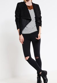 Svart fuskpälsjacka som hänger över en randig topp med bruna detaljer, kombinerad med svarta ripped jeans och ankelstövletter.