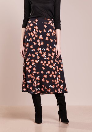 Frau trägt einen schwarzen Midirock mit orange-weißem Blumenmuster, kombiniert mit schwarzen Stiefeln mit Absatz und einem schwarzen Langarmoberteil.