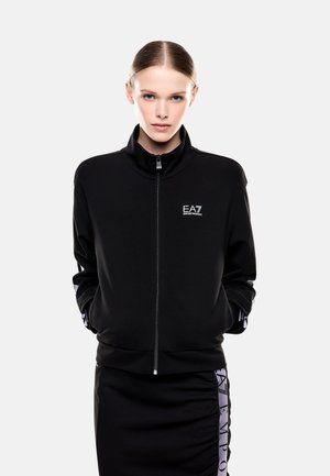 Donna con i capelli pettinati all'indietro che indossa una giacca nera con zip con il logo EA7 Emporio Armani sul petto e una gonna nera abbinata con striscia logo.