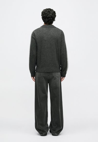 Donkergrijze gebreide trui met een geribde onderrand en kraag, voorzien van witte gestreepte accenten langs de zijkanten. Gecombineerd met losse grijze jeans.