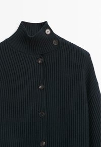 Schwarze, gerippte Strickjacke mit hohem Kragen, die fünf schwarze Knöpfe aufweist, davon zwei am Kragen befestigt und drei vorne entlang.