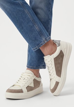 Baskets en daim blanc et beige avec lacets, portées avec un jean en denim bleu, présentées sur un fond clair uni.