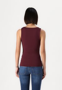 Un débardeur bordeaux avec un col rond et une coupe ajustée, fabriqué à partir de tissu extensible. Porté avec un jean en denim bleu, montrant une texture lisse.