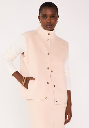 Femme portant un gilet sans manches boutonné rose pâle et une jupe assortie, avec un haut à manches longues blanc et des bijoux en or sur un fond blanc.