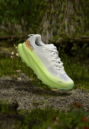 Chaussure de course Hoka blanche et vert citron suspendue au-dessus d’un sol forestier rocheux et moussu.