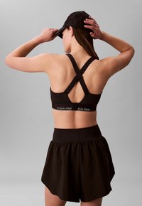 Femeie purtând un sutien sport negru cu bretele încrucișate și o fustă neagră, ajustând o șapcă de baseball neagră, cu spatele spre noi, pe un fundal gri.