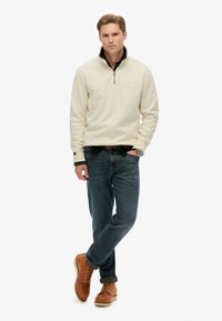 Beige fleece pullover met een kwart ritssluiting, donkere kraag, blauwe spijkerbroek en beige veterschoenen. De outfit heeft een gelaagde look met een zwarte shirt eronder.