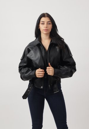 Donna con lunghi capelli scuri che indossa una giacca di pelle nera e jeans scuri, che tiene il colletto della giacca, in piedi contro uno sfondo chiaro e uniforme.