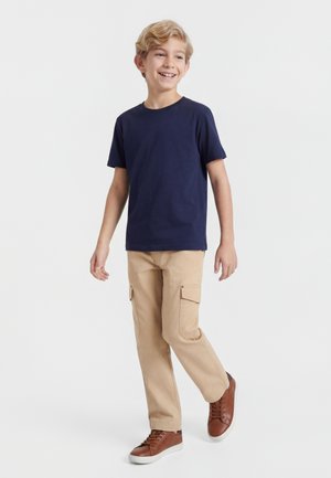 Usmievajúci sa blondiak chlapec kráča, oblečený v námorníckom modrom tričku, béžových cargo nohaviciach a hnedej športovej obuvi na jednofarebnom svetlom pozadí.