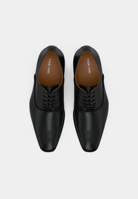 Černé kožené společenské boty s špičatou špičkou, které mají hladký povrch, minimalistický design a hnědé vnitřní stélky s logem značky.