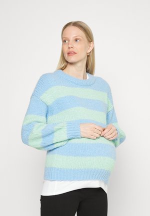 Suéter oversized em azul claro e verde menta com riscas, com um decote redondo e mangas largas, usado sobre uma blusa branca e calças pretas.