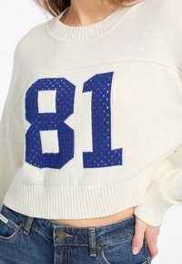 Pull en tricot blanc avec le numéro bleu "81" en texture ajourée, avec un col et des poignets côtelés, associé à un jean en denim bleu.
