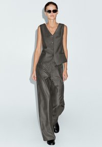 Donna che cammina con un gilet senza maniche a righe e pantaloni larghi abbinati, con scarpe nere e occhiali da sole scuri, su uno sfondo neutro.