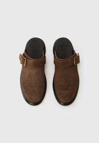 Zoccoli in suede marrone con una larga fascia e una fibbia in ottone, superficie testurizzata e suola in gomma nera, vista dall'alto.