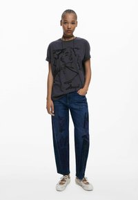 T-shirt en coton gris foncé avec un motif graphique, associé à un jean bleu ample avec des illustrations violettes foncées, et des baskets rétro blanches.