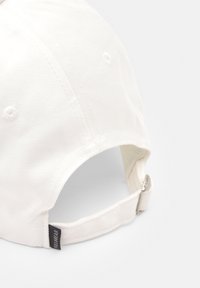 Casquette de baseball en coton blanc avec une visière curved, sangle réglable à l'arrière, et une petite étiquette de marque attachée sur le côté.
