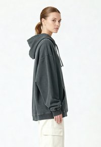 Felpa con cappuccio oversize di colore grigio scuro, materiale in cotone, tasca a marsupio, cappuccio con coulisse, polsini e orlo a coste. Indossata con pantaloni cargo chiari.