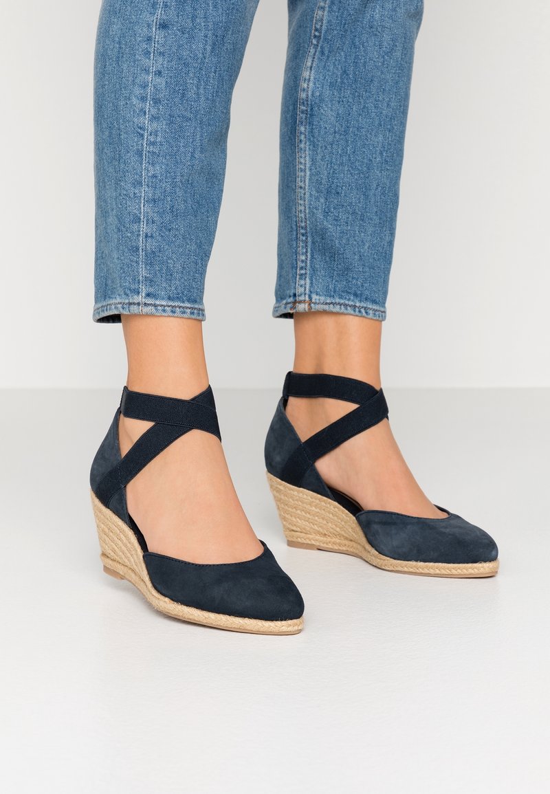 Pieds portant des espadrilles compensées en daim bleu marine avec des brides croisées autour de la cheville, associées à un jean bleu raccourci.