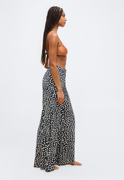 Moteris su ilgomis kasomis, dėvinti rudos spalvos bikini viršutinę dalį ir juodas, plačias, aukštu liemeniu kelnes su baltomis taškelėmis, stovinti basomis šonu.