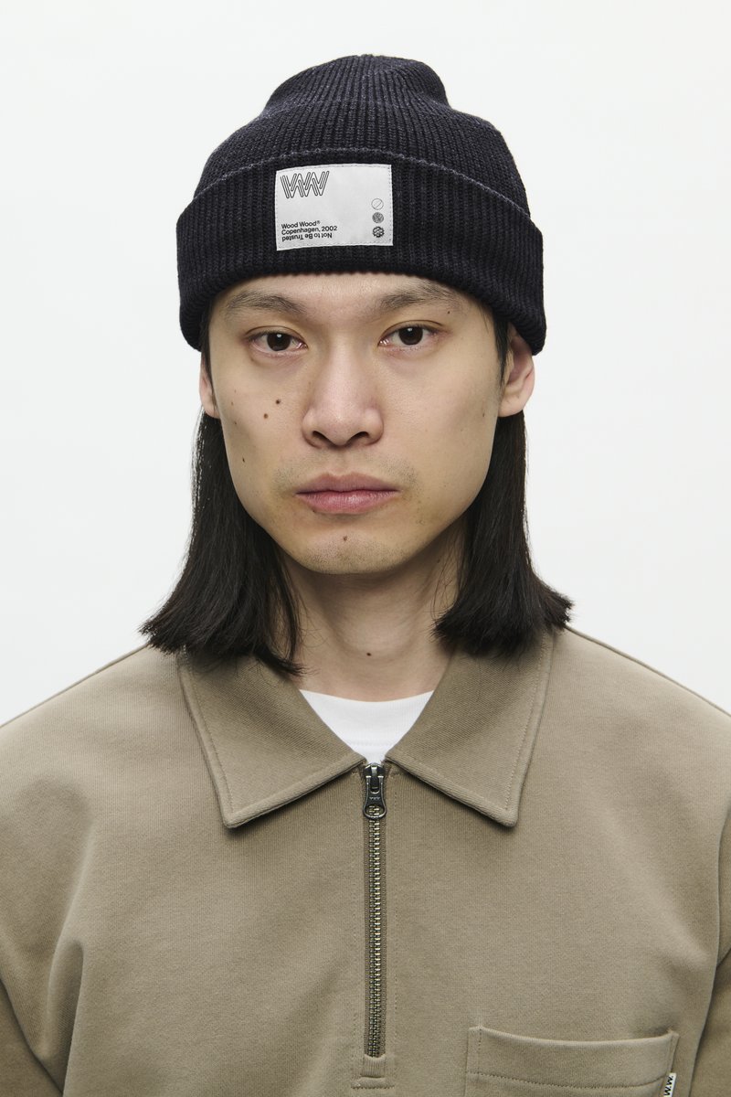 Man met lang zwart haar draagt een zwarte gebreide muts met een wit label en een beige jas met ritssluiting over een wit shirt.