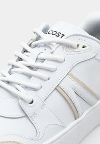 Gros plan d'une basket blanche avec des lacets blancs et des accents beiges subtils, montrant une partie du nom de la marque sur la languette.