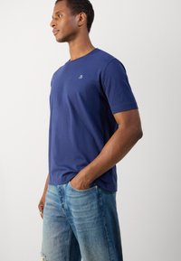 Mørkeblå kortærmet t-shirt i blødt materiale, med en rund halsudskæring og et lille logo på brystet. Sammen med lyseblå slidte jeans.
