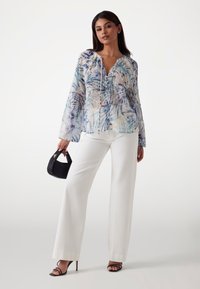Bluză transparentă cu model floral, accentuată cu nuanțe de albastru și verde, cu guler strâns și mâneci lungi, asortată cu pantaloni largi albi și o geantă neagră.