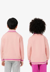 Růžová mikina s žebrovaným límcem a manžetami, která má tmavě růžové pruhy. Je oblečená s šedými tepláky a růžovými kalhotami. Pohled zezadu.