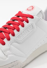 Sneaker bianco con lacci rossi, tomaia in pelle testurizzata, logo adidas impresso e dettagli perforati sui lati. Suola in gomma robusta.