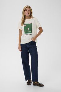 Camiseta crema con gráfico temático de tenis en verde y rosa, que presenta el texto "COURT SPORT". Llevada con jeans oscuros y holgados y mocasines marrones.