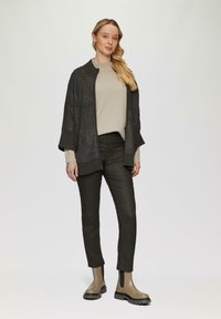 Grau gestrickte Strickjacke mit strukturierten Ärmeln, beigefarbenem Pullover darunter, dunklen Hosen und beigefarbenen Stiefeletten mit dicker schwarzer Sohle.