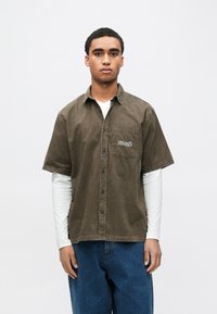 Camisa castanha de manga curta com botões em tecido texturizado, com um bolso no peito e desenho colorido bordado, usada sobre uma camisa branca de manga comprida.