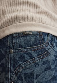 Gros plan d'une poche de jean en denim bleu à motif avec des coutures orange et un ourlet de pull en maille beige.