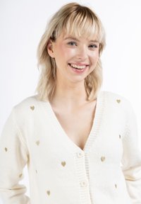 Bílý cardigan vyrobený z měkkého materiálu, s zlatými srdíčky, V-výstřihem a třemi knoflíky na přední straně.
