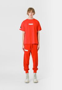 Camiseta corta de algodón rojo con un parche rectangular blanco, combinada con pantalones a juego que tienen bolsillos laterales y detalles de flecos, y botas blancas.