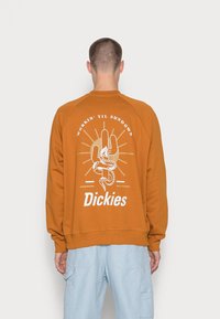 Oranžová mikina s kulatým výstřihem má na sobě grafiku ruky a hada v bílé barvě, s nápisem "Pracuji až do západu slunce" nahoře a "Dickies" dole.