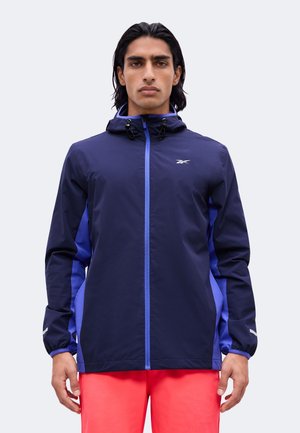 Homme portant une veste à capuche bleu marine et violette avec une fermeture éclair frontale et un pantalon rouge vif, debout devant un fond uni.