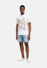 Camiseta de algodón blanca con estampado floral y texto en azul y dorado, combinada con pantalones cortos de mezclilla azul claro y zapatillas blancas.