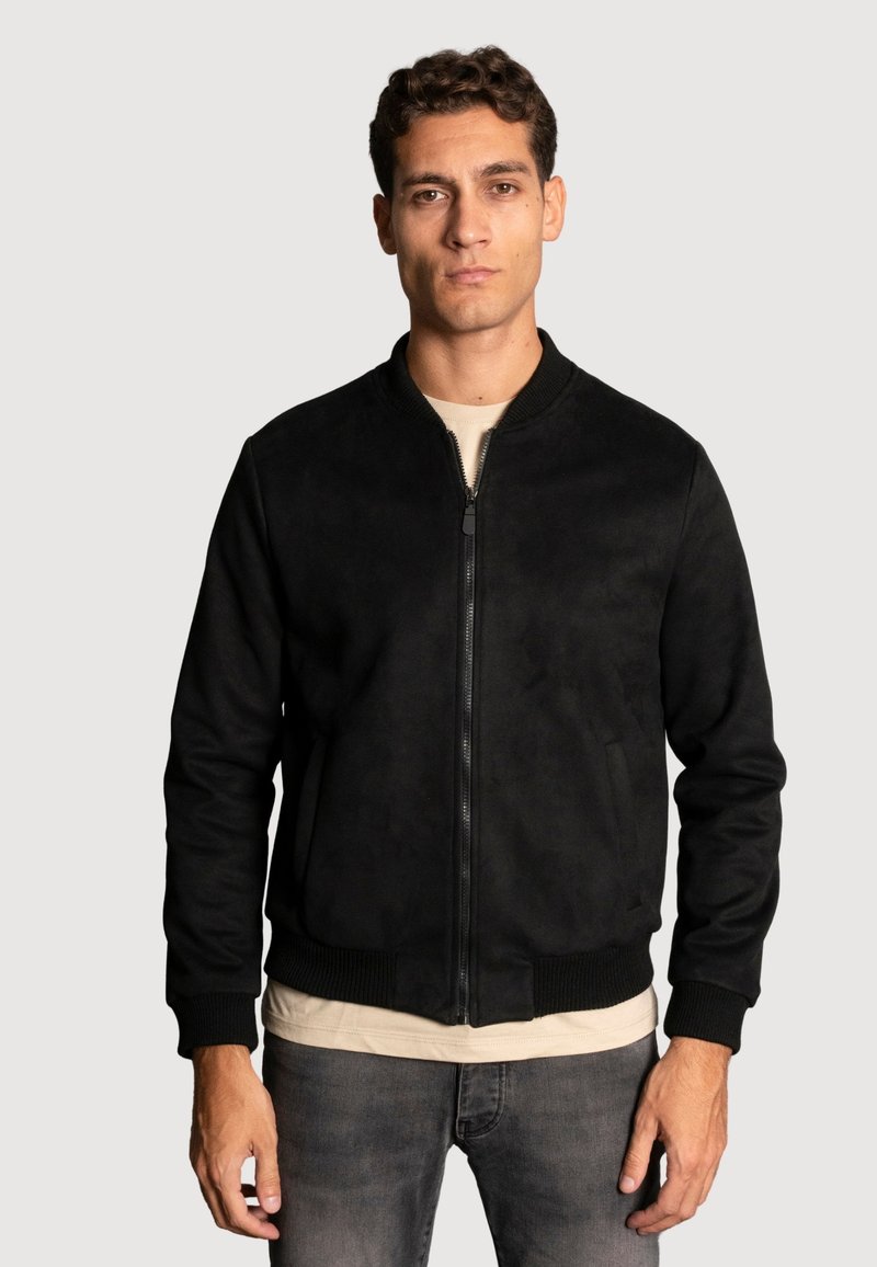 Veste bomber noire en tissu doux, avec fermeture zippée, poignets et taille côtelés, portée par-dessus une chemise beige, sur fond uni.