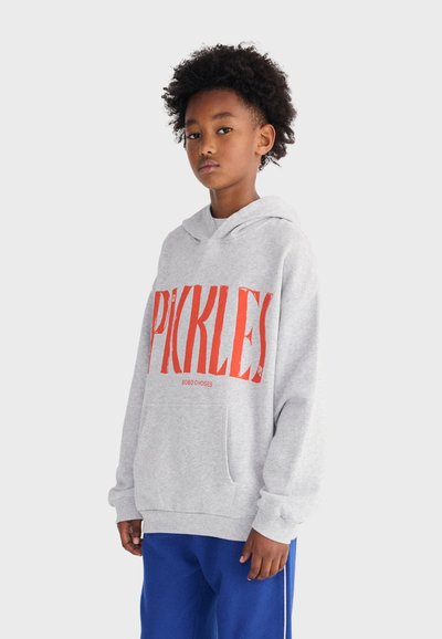 Niño con cabello rizado que lleva una sudadera con capucha gris claro con la palabra "PICKLES" en grandes letras rojas y pantalones azules, de pie contra un fondo liso.