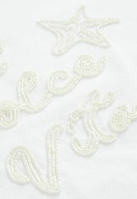Tissu blanc présentant une broderie en relief, perlée, dans un style cursif et en forme d'étoile, mettant en avant un design texturé et complexe.