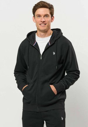 Junger Mann lächelt, trägt schwarzen Kapuzenpullover mit Reißverschluss und passende schwarze Hose mit kleinem weißem Logo, Hände in den Taschen des Kapuzenpullovers, weißes Hemd darunter.