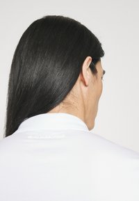 Hvit polo-shirt med krage, laget av myk stoff og med en subtil preget logo på baksiden. Lang, rett mørk hår synlig.