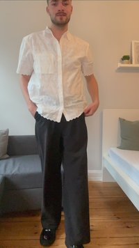 Camicia bianca in pizzo con bottoni, maniche corte, orlo scallop e una tasca, abbinata a pantaloni neri a gamba larga e scarpe nere.