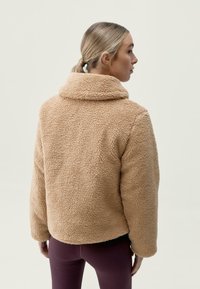 Beige, huňatý fleecový kabát s vysokým límcem, uvolněným střihem a dlouhými rukávy. Nosený přes tmavě fialové legíny. Viditelná strukturovaná tkanina.