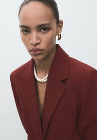 Jonge vrouw in een bordeauxrode blazer met een parelketting en gouden oorbellen tegen een effen lichte achtergrond.