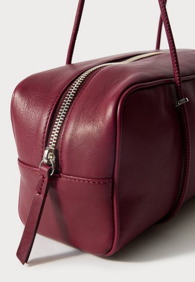 Burgundin värinen nahkalaukku, jossa on suorakulmainen muoto, hopeinen vetoketjusuljenta ja lyhyt nahkahihna. Sileä pinta ja siistit tikkaukset.